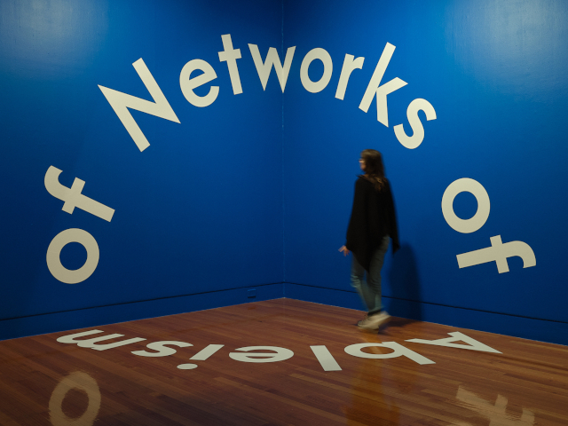 Two deep blue walls meet at the corner of a high room with a wooden floor. Large white lettering runs in a circles across the walls and over the floor with the words 'Ableism of Networks of' formign a never-ending sentence. The circle of letters is larger than the height of a person. A woman stands inside the letterng circle to one side of the corner.