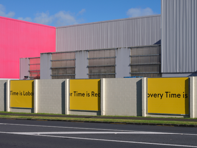 A set of three billboard posters mounted on panels running along the wall of a modern industrial looking building. The posters are plain yellow with black lettering that runs across the middle of all thre boards, reading: Time is Labour Time is Recovery Time is